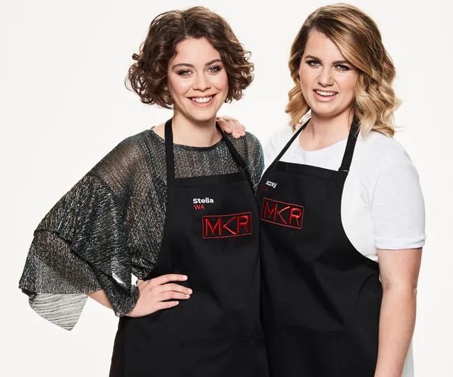 Two women wearing "MKR" aprons stand together smiling at the camera against a plain background.
