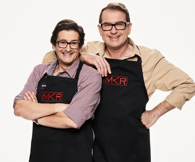Two men in "MKR" aprons smiling, one with arm around the other, against a plain background.