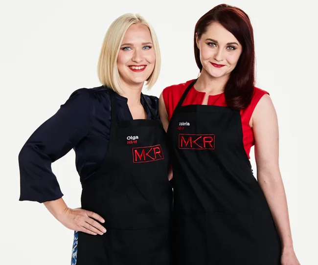 Two women in "My Kitchen Rules" aprons, smiling, one with blonde hair and one with dark hair, on a white background.