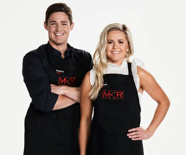 Two contestants from "My Kitchen Rules" wearing black aprons, smiling. One has "Henry" and the other has "Anna" on their aprons.