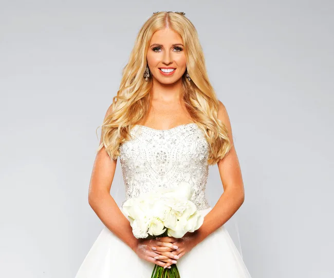 A woman in a white wedding gown holds a bouquet of white roses against a light gray background.