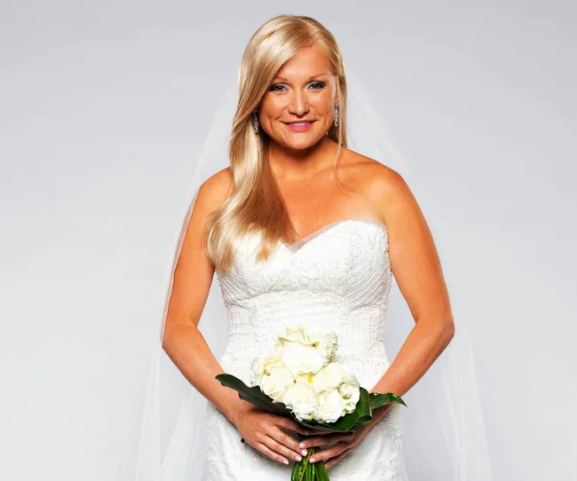 Blonde woman in a strapless wedding dress holding a bouquet of white roses, smiling.