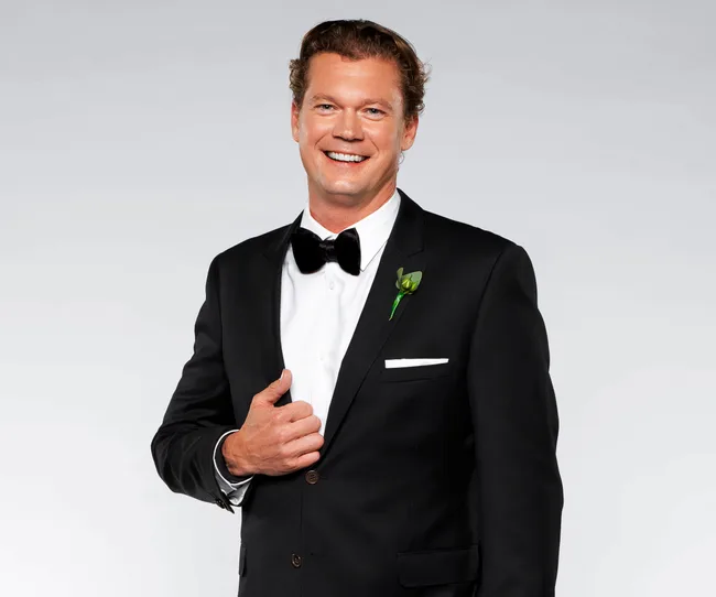 Man in a black suit with a bow tie, holding lapel, smiling against a gray background.