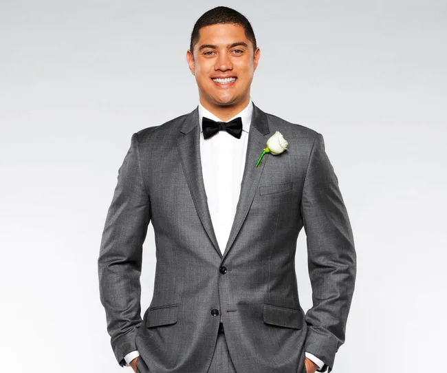 Man in a gray suit with a bow tie and white rose boutonniere, smiling against a plain backdrop.