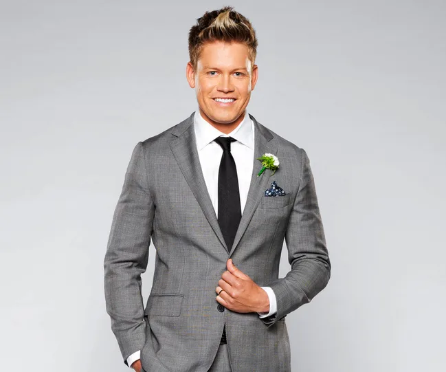 Man in gray suit smiling, wearing a black tie, with a boutonniere and pocket square against a plain background.