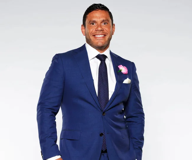 Man in a blue suit with a pink boutonniere, smiling against a plain background.