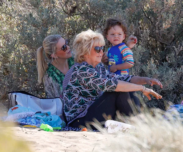 Three people sitting outdoors on sand, including a woman with sunglasses and a child, surrounded by bushes.