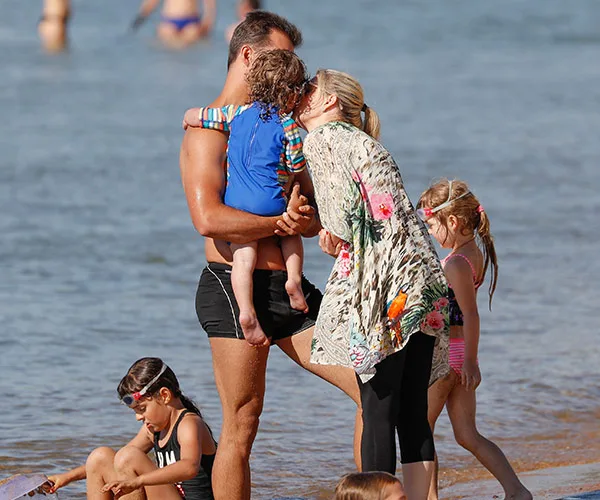Family enjoying beach day; woman kisses man holding child, surrounded by kids playing in sand and water.