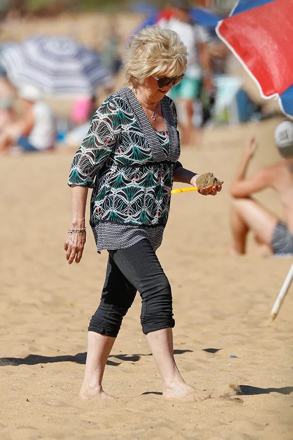 A woman in sunglasses walks on a sandy beach, holding a stone and a yellow object.