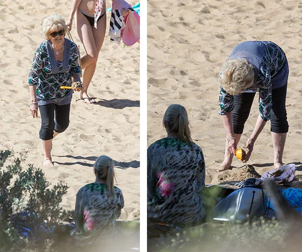 Older woman walking on beach holding toy; then crouched near seated person, playing with sand.