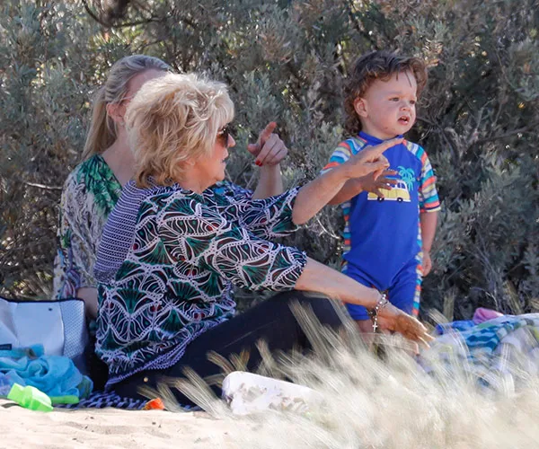 Three people, including an older woman and a child in a blue outfit, seated on a sandy beach with trees in the background.