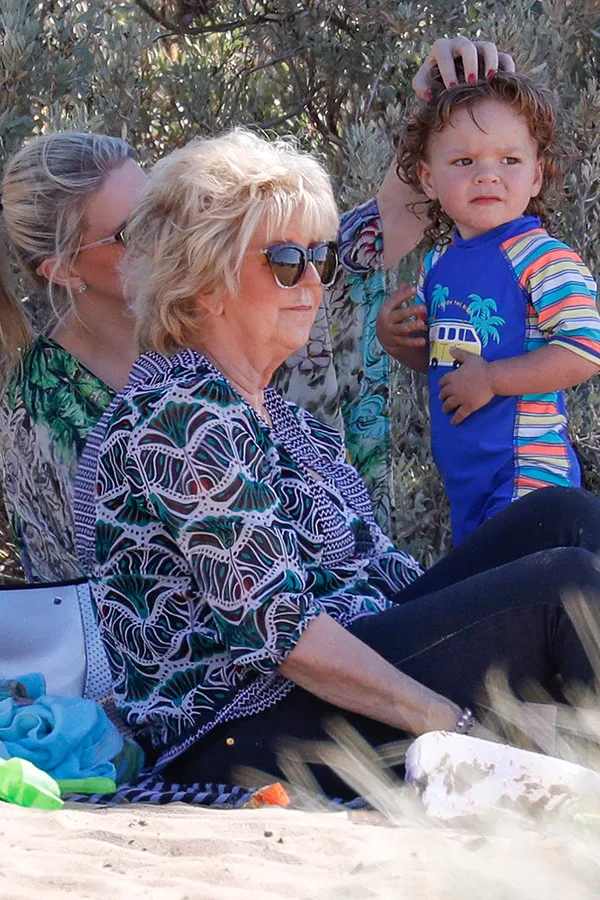 Three people on a beach; a woman wearing sunglasses and a patterned shirt is seated, with a child in a colorful outfit standing beside her.