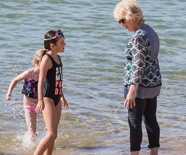 Adult talking with two children playing in the water at a beach, one wearing a black swimsuit with text.