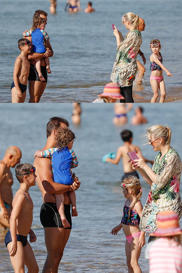 Beach scene with adults and children playing in the water, woman taking photos with a smartphone.