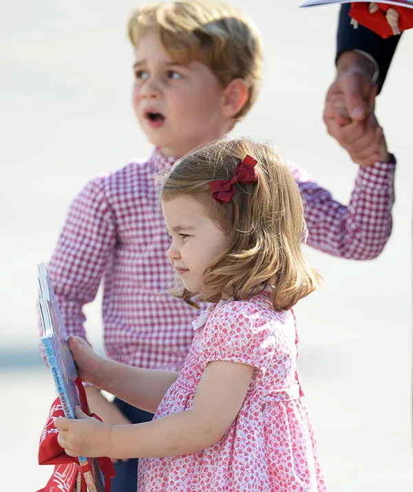 Children holding hands, one holding papers, dressed in checkered and floral outfits.
