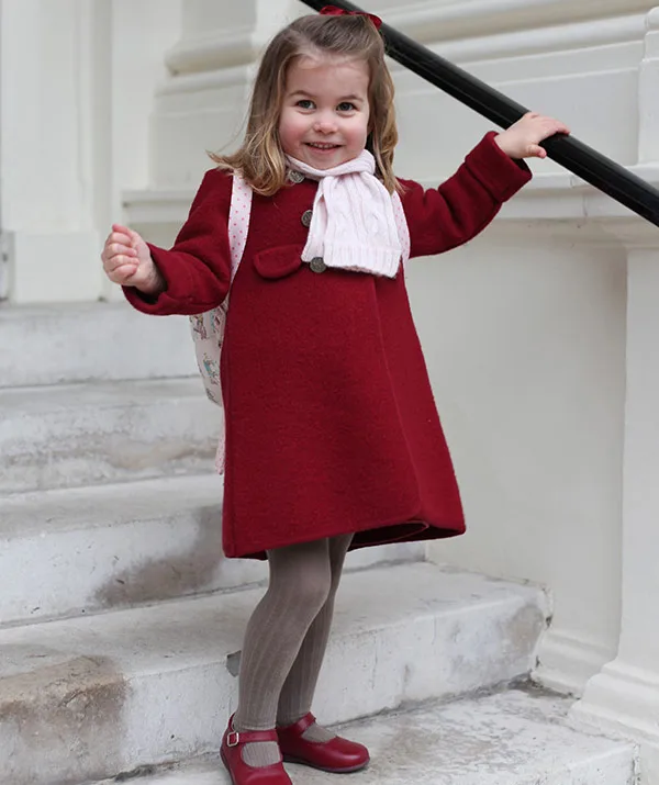 A young girl in a red coat, tights, and a scarf smiles on stone steps, holding a railing.