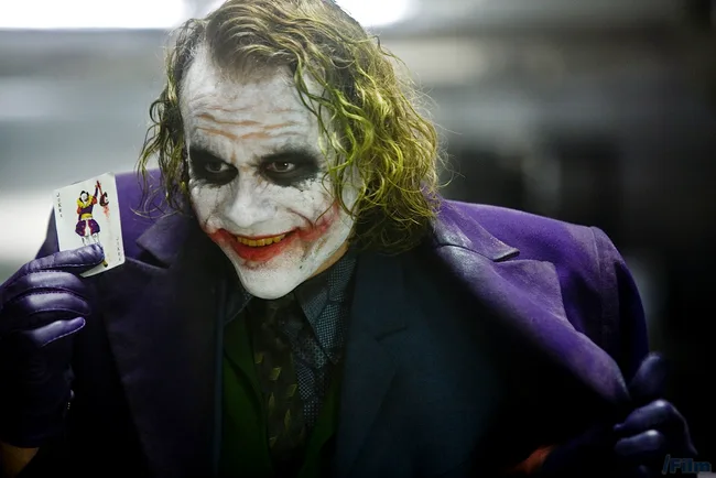The Joker from The Dark Knight holding a Joker playing card, wearing a purple suit and gloves, with a sinister smile.
