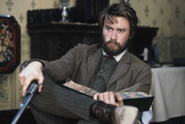 A bearded man in period attire holds a rifle and a book, sitting in a vintage room with patterned wallpaper.