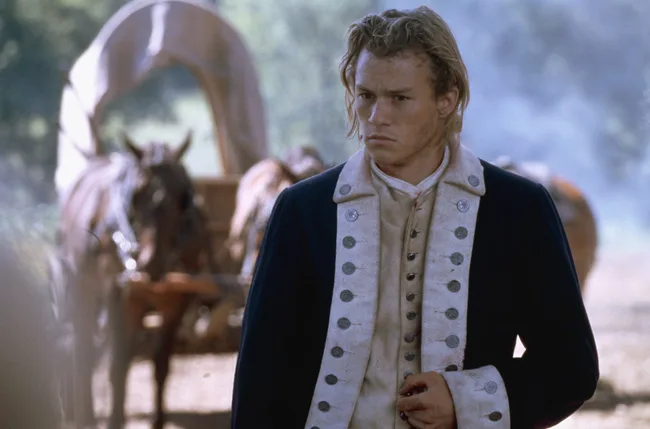 A young man in a Revolutionary War uniform stands in front of a blurred covered wagon and horses.