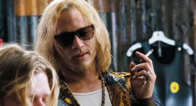 A man with sunglasses, long blond hair, and a cigarette, wearing a patterned shirt and beads, in a casual setting.