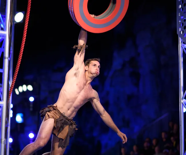 Competitor swings on a giant ring, dressed in a loincloth, under dramatic blue lighting on an obstacle course.