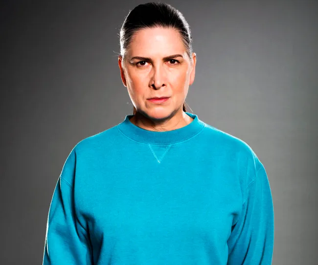 Woman in a teal sweatshirt with a serious expression against a gray background.