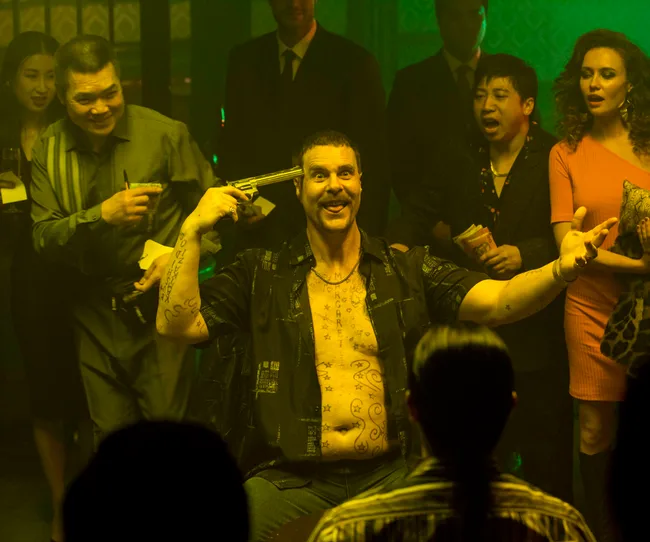 A man with tattoos holds a gun to his head while surrounded by a crowd in a dimly lit room, creating a tense atmosphere.