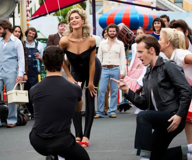 A woman in black attire poses confidently as two kneeling men in leather jackets point and sing, surrounded by a cheering crowd.