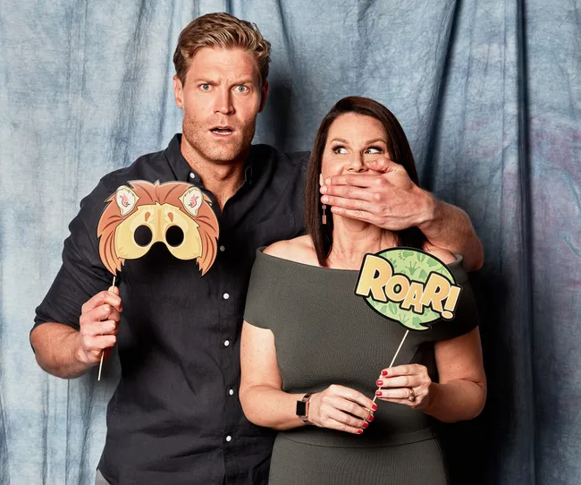 Two people posing with props; the man holds a lion mask, and the woman has a "Roar!" sign, his hand covers her mouth.