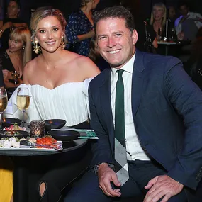Two people seated at a table with drinks and food, smiling at a social event.