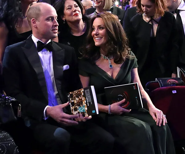 A man in a tuxedo and a woman in a green dress holding programs, seated at an event with people in the background.