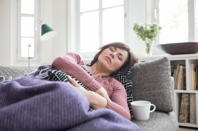 Woman resting on a sofa with a cup nearby, wrapped in a purple blanket, in a bright living room.