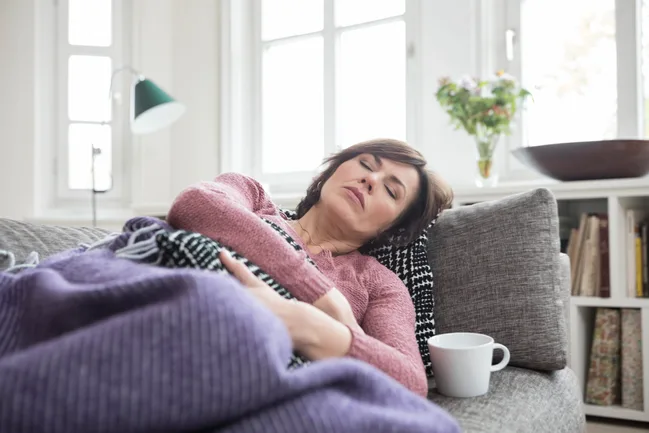 Woman resting on a sofa with a cup nearby, wrapped in a purple blanket, in a bright living room.