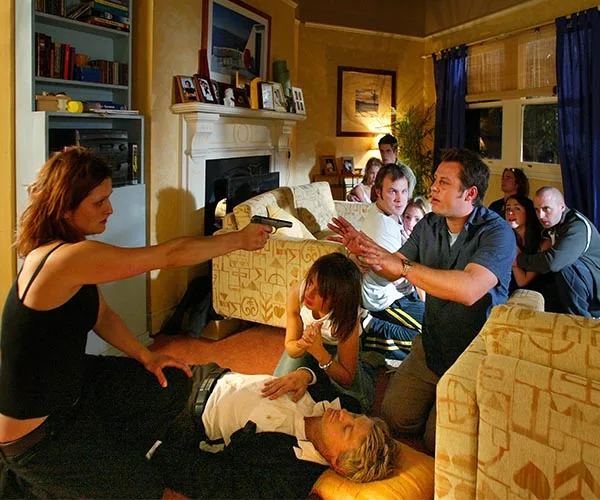 A tense living room scene with a woman holding a gun, pointing it at a group of people with hands raised, one person on the floor.