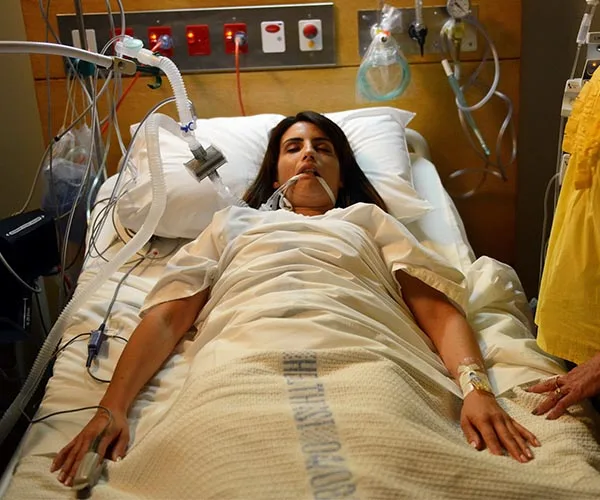 A woman lies unconscious on a hospital bed, connected to medical equipment and tubes.