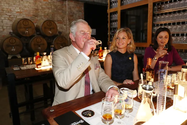 Man in beige suit tasting whisky at a bar with two women, barrels and glassware in the background.