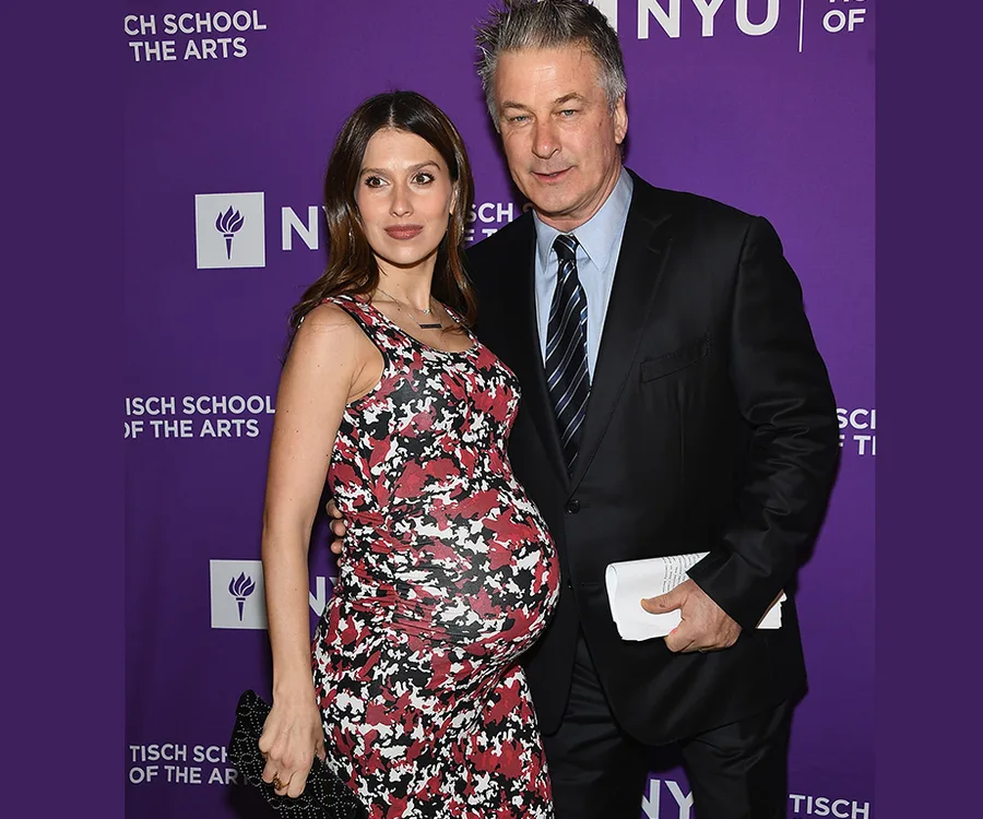 A couple poses on a red carpet, the woman is visibly pregnant in a floral dress, and the man wears a suit and tie.