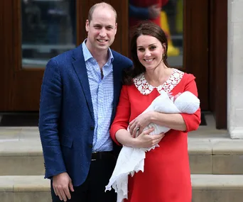 A couple poses joyfully on steps, the woman cradling a newborn wrapped in a white blanket.