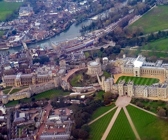 Aerial view of Windsor Castle surrounded by green lawns and trees, with the River Thames in the background.