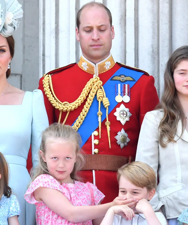Royal family members posing, with a young girl playfully covering a boy's mouth, a man in a red uniform stands behind.