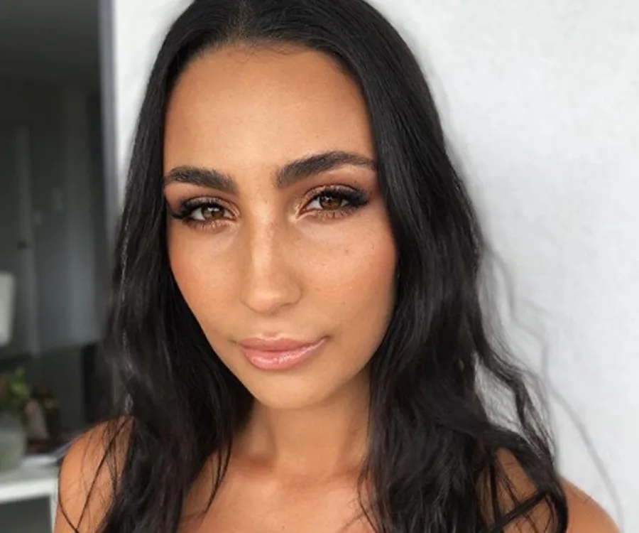 A woman with long dark hair and eyelash extensions, wearing natural makeup, posing indoors.