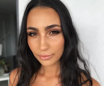 A woman with long dark hair and eyelash extensions, wearing natural makeup, posing indoors.
