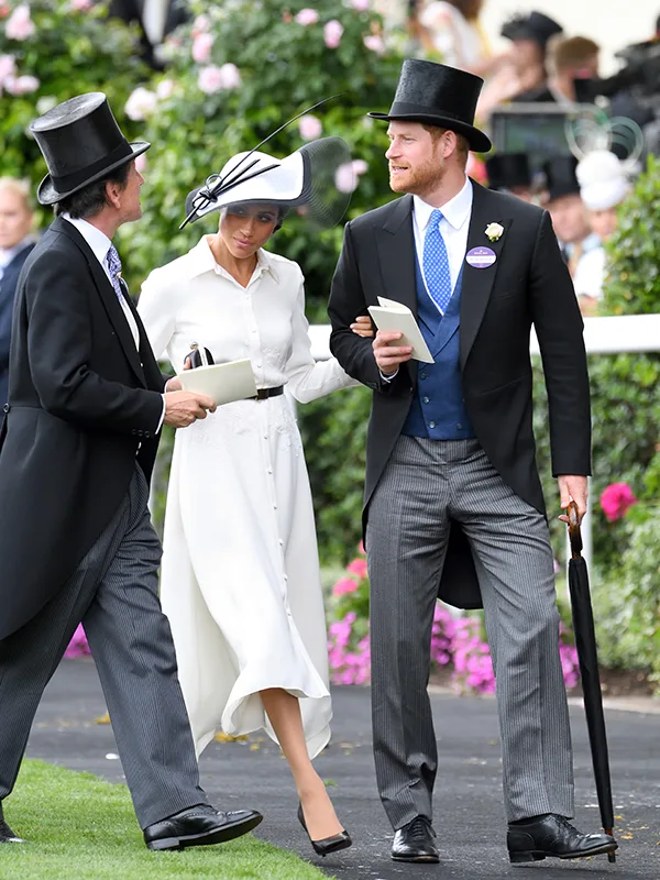 Couple in formal attire with top hats and umbrella, walking and smiling at Royal Ascot event.
