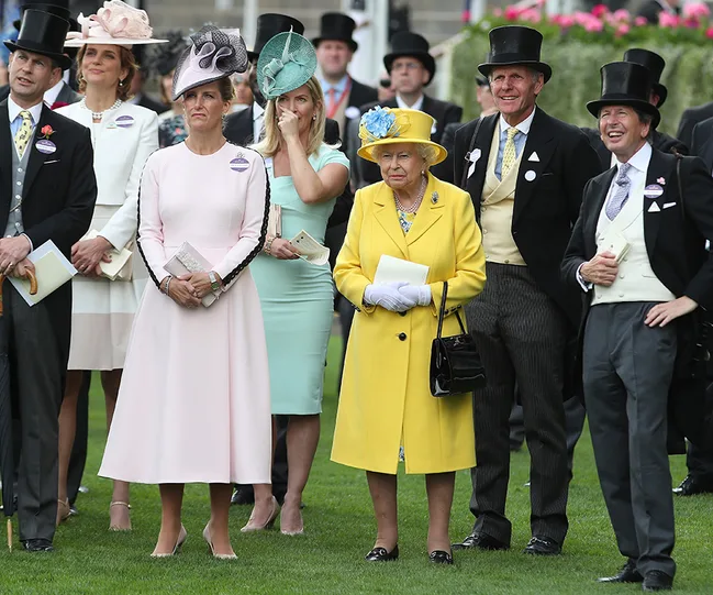 A group of people in formal attire, with hats, stand at Royal Ascot on a grassy area.