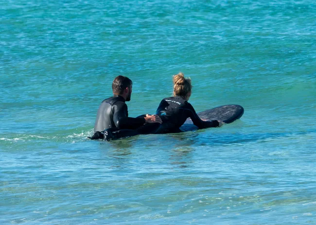 Two people in wetsuits on a surfboard, paddling in calm ocean water.