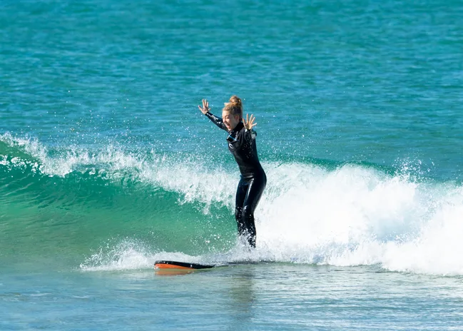 "Person in black wetsuit surfing a small wave, arms raised in excitement on a sunny day."