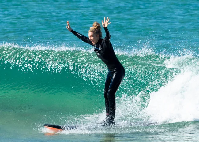 Person surfing on small wave, wearing wetsuit, hands raised.