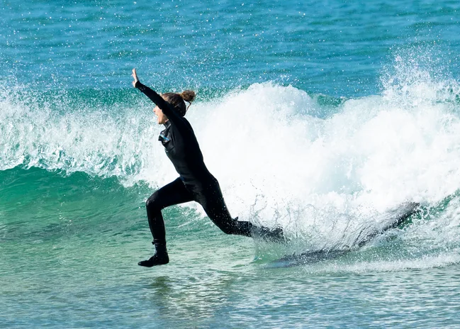 Surfer in a wetsuit leaping off a wave with arms raised, ocean in the background.