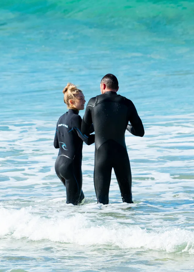 Two people in wetsuits standing in shallow ocean water.
