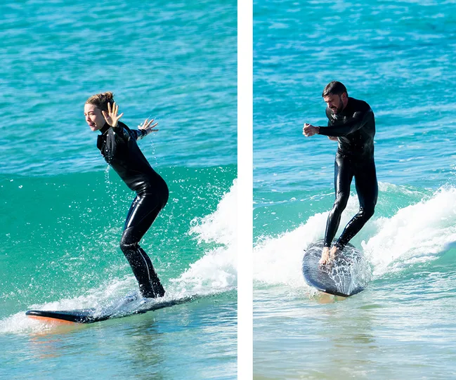 Two people surfing on small waves, both wearing black wetsuits, against a backdrop of blue-green ocean water.